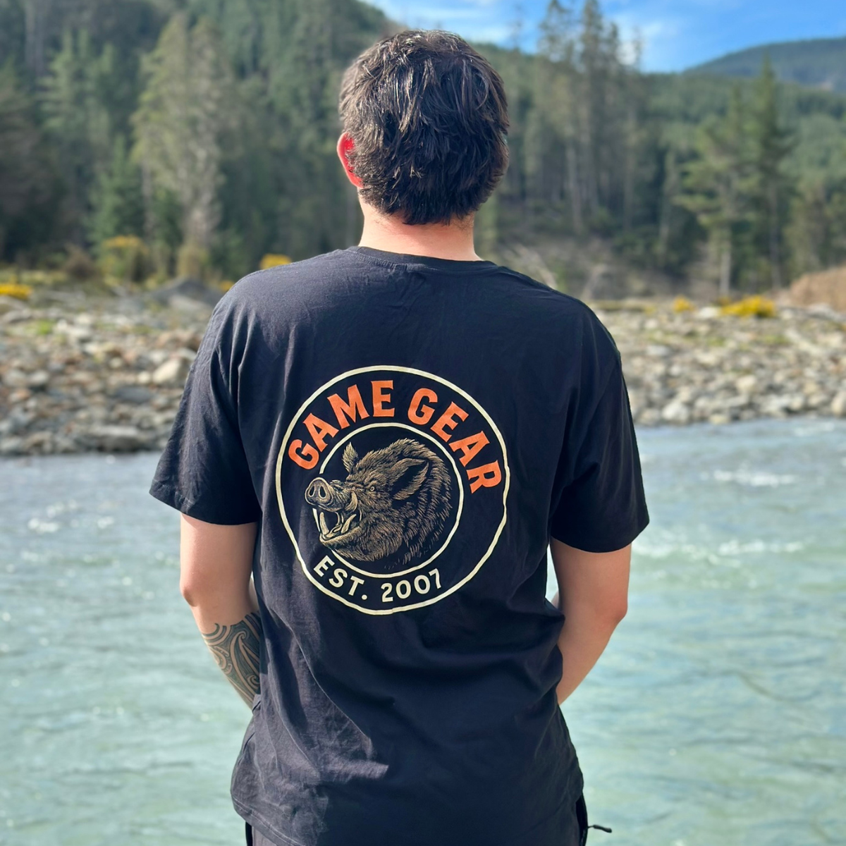 Person wearing a black t-shirt with 'Game Gear' logo facing a lake with trees in the background