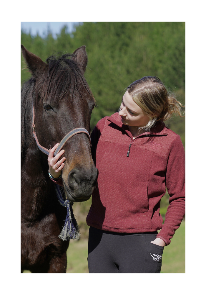 Woman in a red fleece jacket petting a horse outdoors with greenery in the background